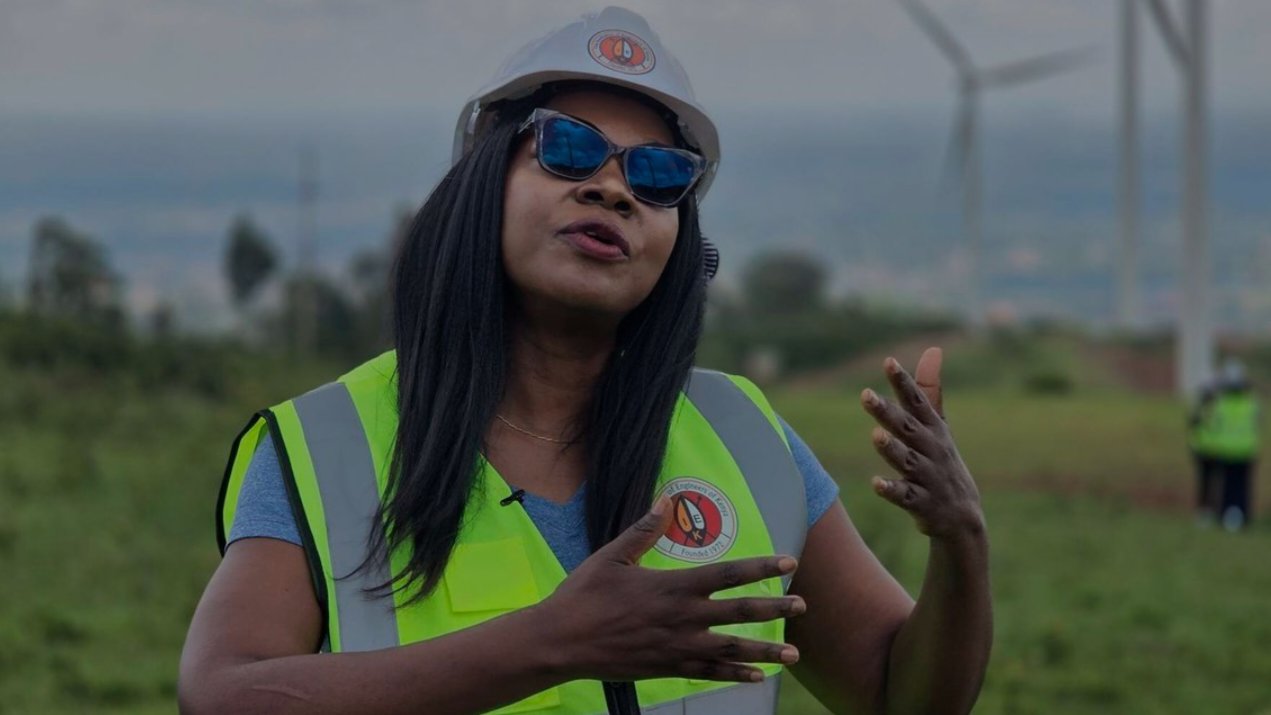Engineer Jennifer Gache wearing a high-visibility safety vest and a white hard hat speaking at an outdoor wind energy installation site with wind turbines in the background.