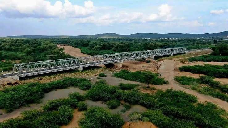An aerial view of the steel truss Kalobeyei bridge spanning a dry riverbed surrounded by green scrubland in Turkana County.