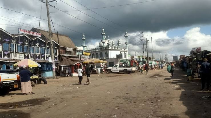 A wide-angle view of a dusty commercial area in Kyumbi showing a mosque, several small shops, parked vehicles, and people walking along the unpaved roadside under a cloudy sky.