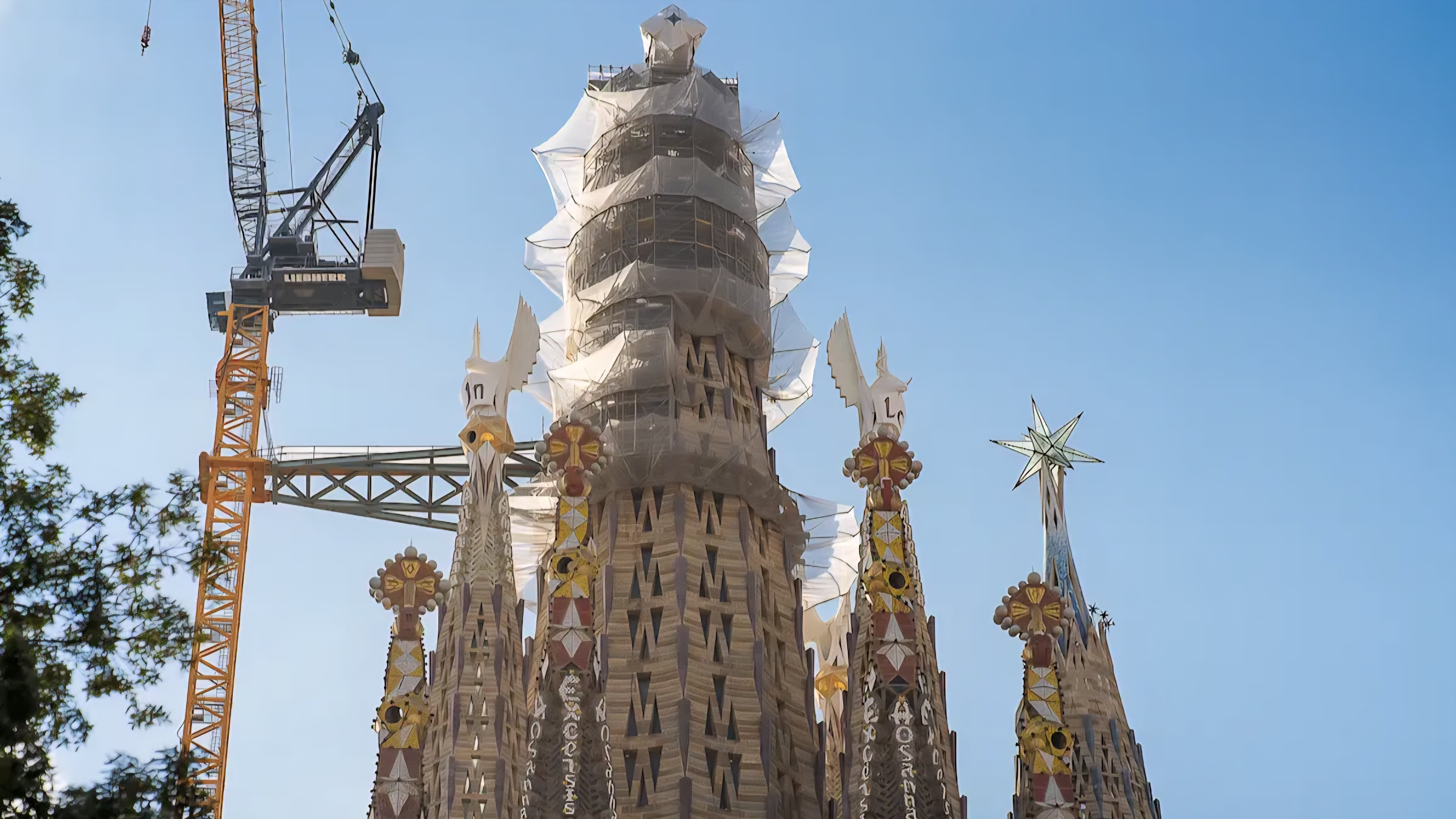An aerial view of the Sagrada Familia basilica in Barcelona showing the newly completed central towers surrounded by cranes under a clear blue sky, as referenced in file 250153.png.