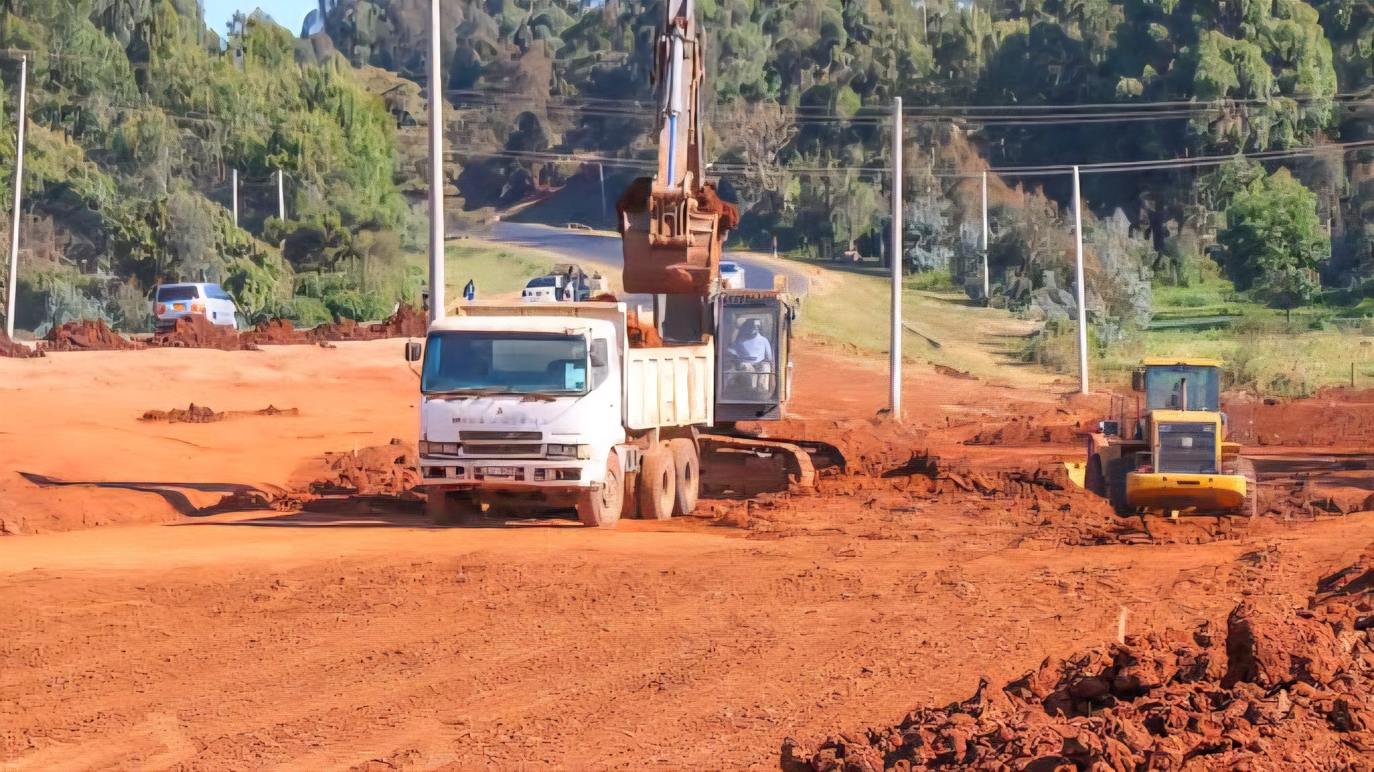 An excavator and a truck working on a dirt section of a road construction site with trees in the background.