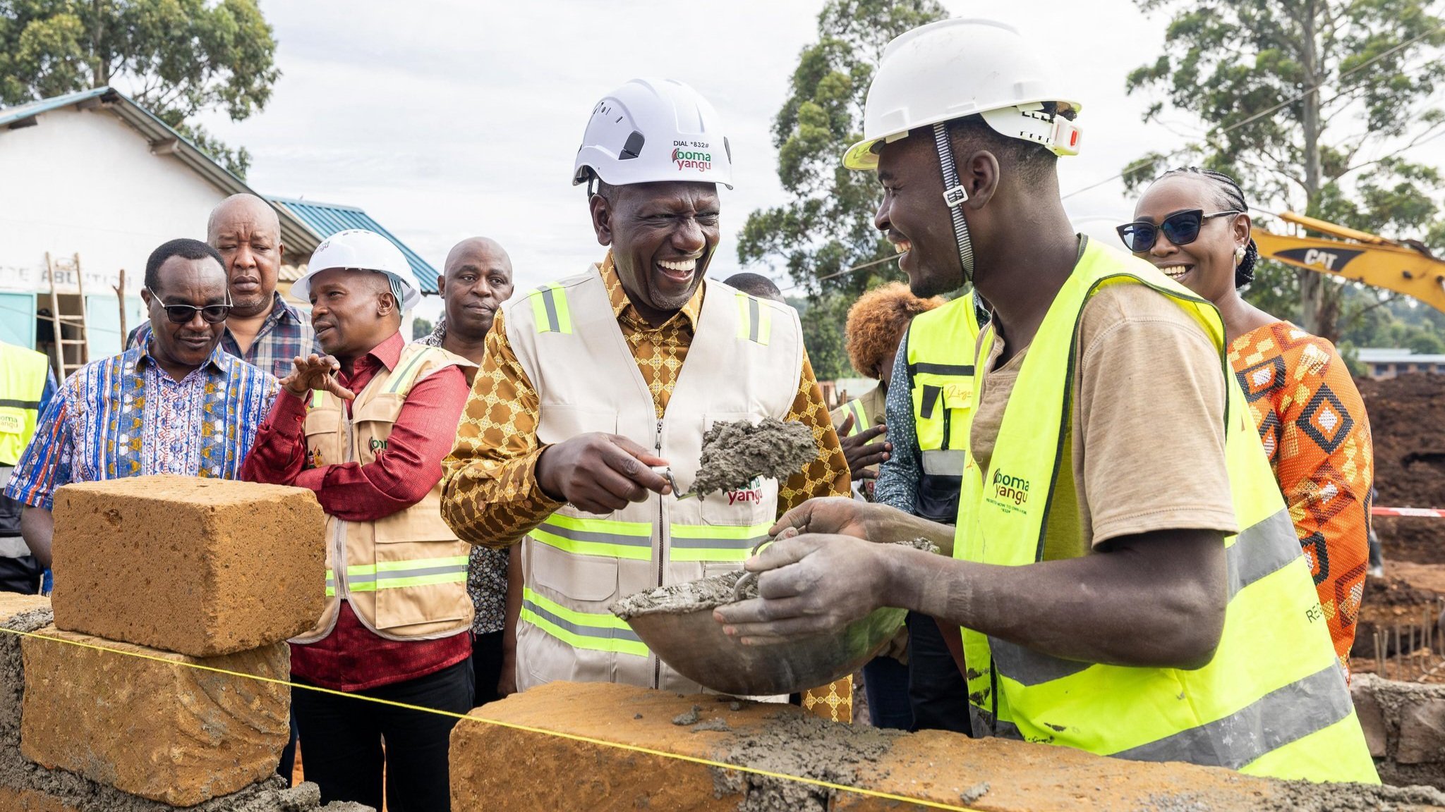 President William Ruto wearing a white hard hat and yellow reflector vest uses a trowel to apply mortar to a stone during a groundbreaking ceremony in Nyamira.