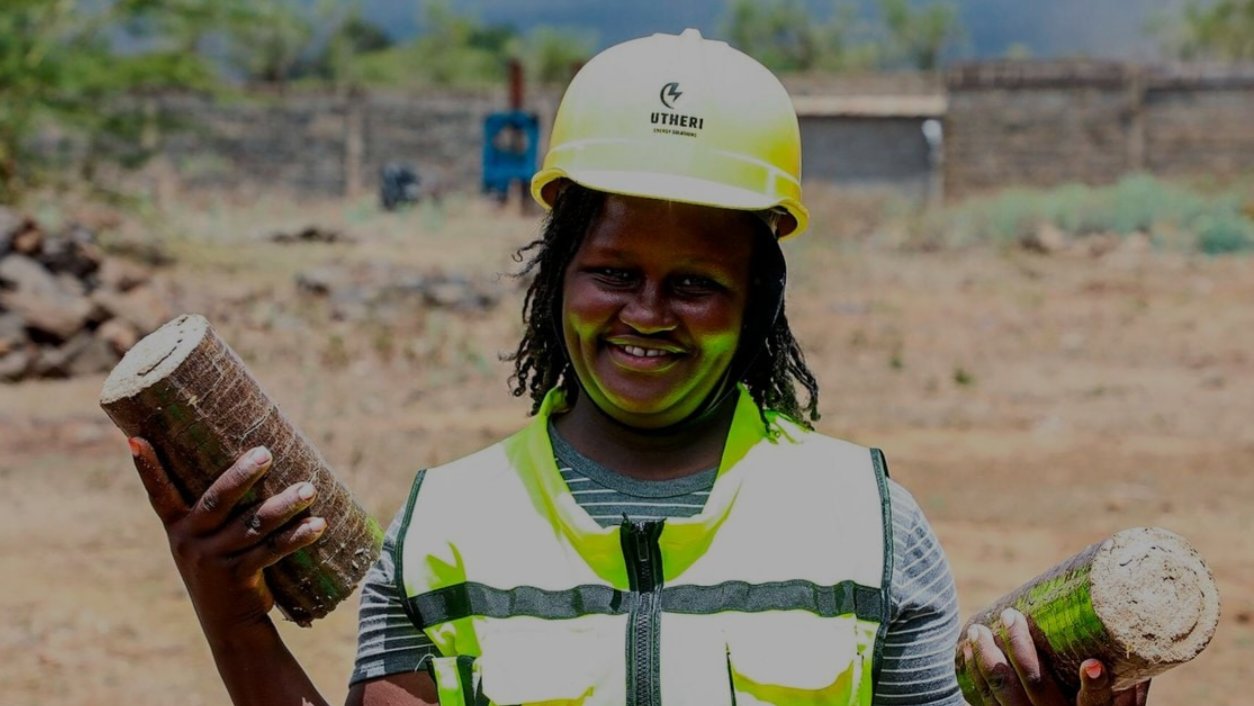 A woman wearing a yellow hard hat and a high-visibility safety vest smiles while standing at a briquette production site.