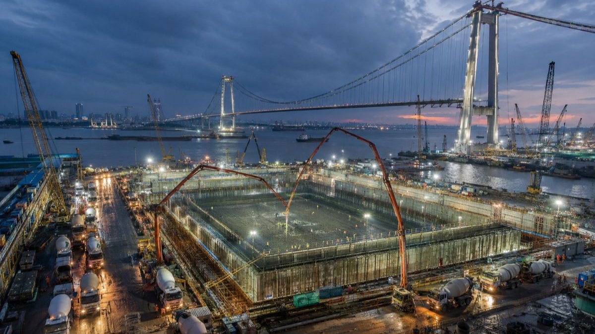 A night view of a brightly lit, massive foundation caisson construction site on the banks of a river with a suspension bridge visible in the background.