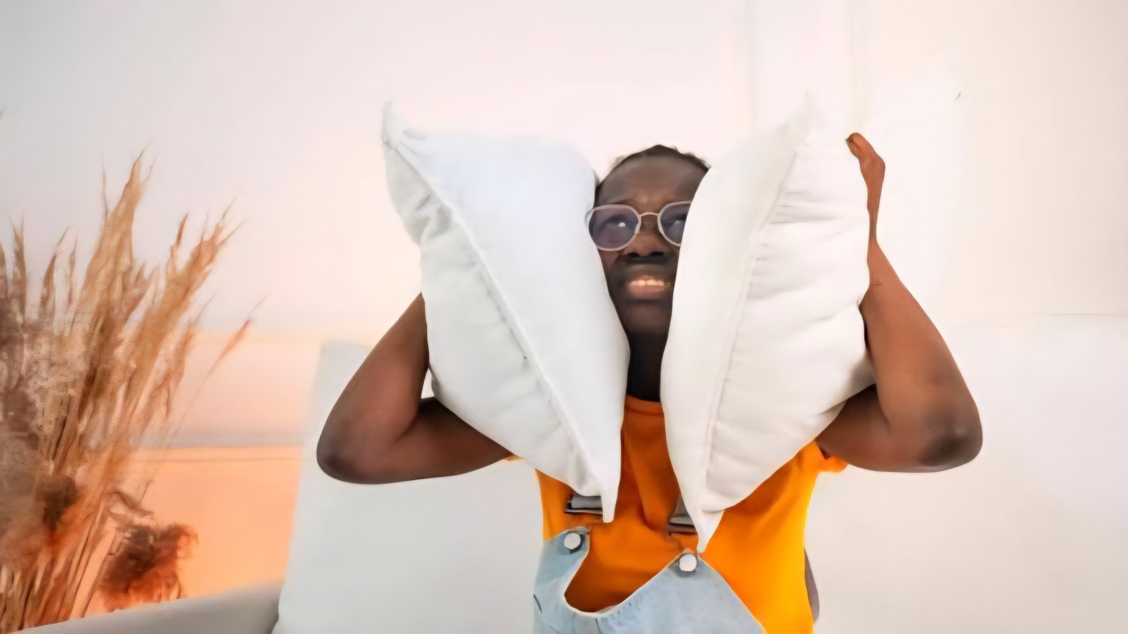 A man covering his ears with white pillows to block out loud noise in a residential setting.