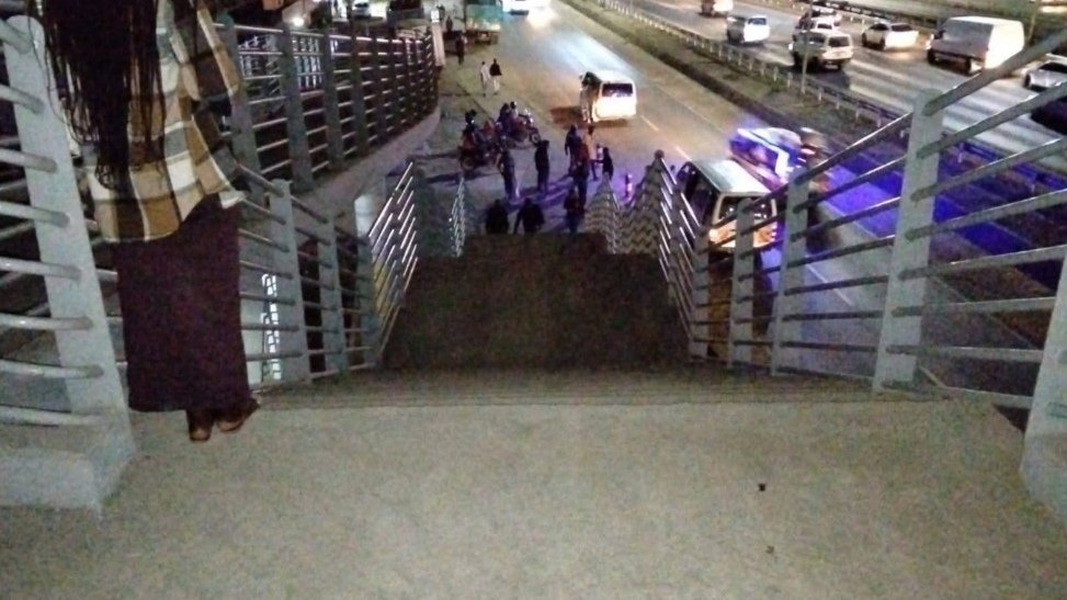 An elevated view of a modern footbridge on a busy highway at dusk. A group of pedestrians walks across the clear, well-lit path while light trails from traffic streak across the road below.