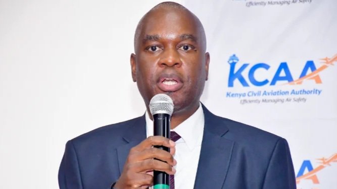 A portrait of a Kenya Civil Aviation Authority official speaking into a microphone with the KCAA logo visible in the background.