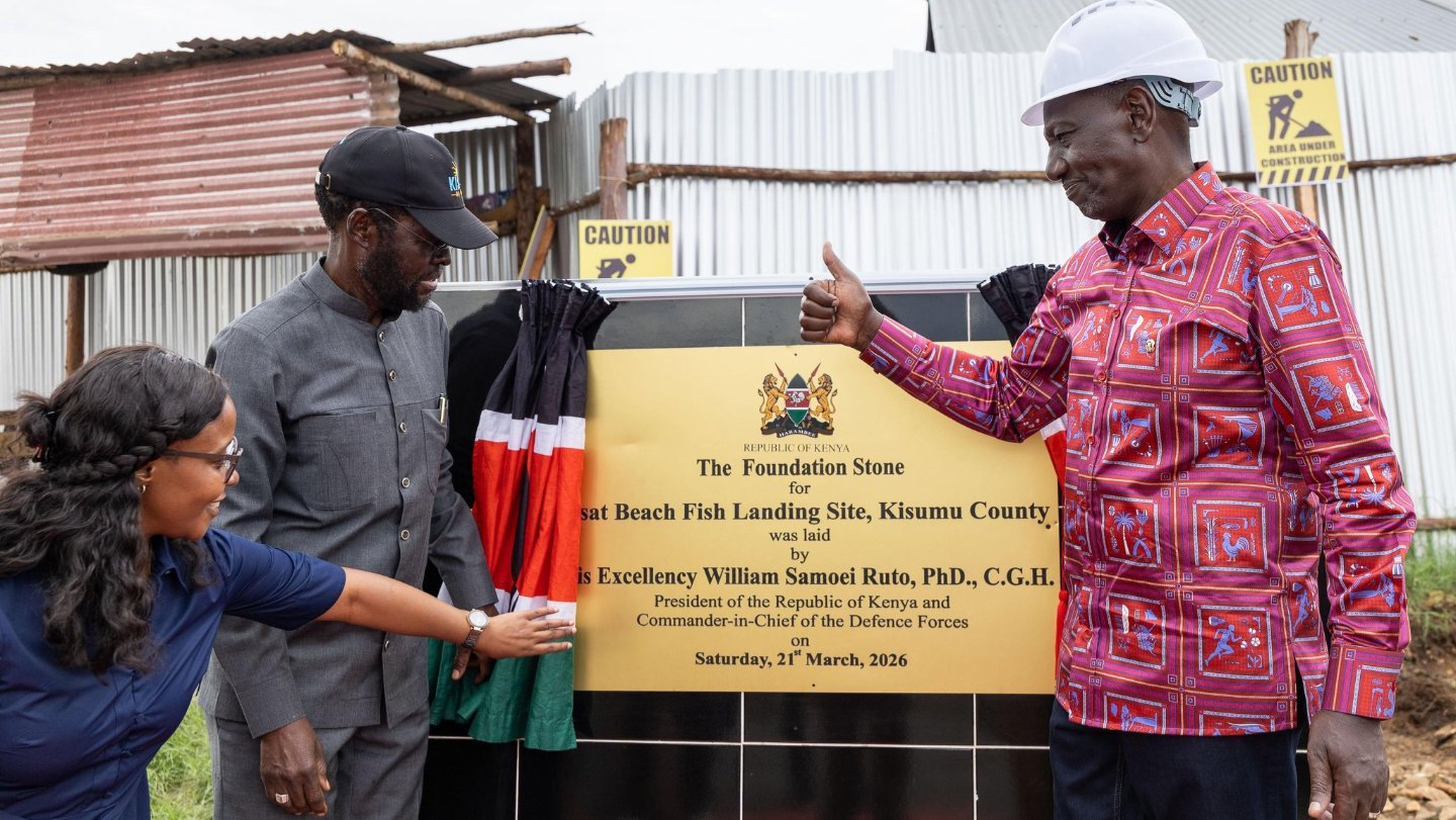 President William Ruto standing with officials at the foundation stone of the Asat Fish Landing Site in Kisumu, with construction work and a large crowd visible in the background.