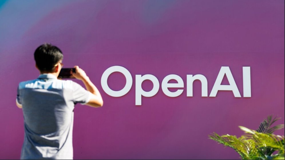 A person holding a mobile phone taking a photograph of an OpenAI signage setup against a gradient background.