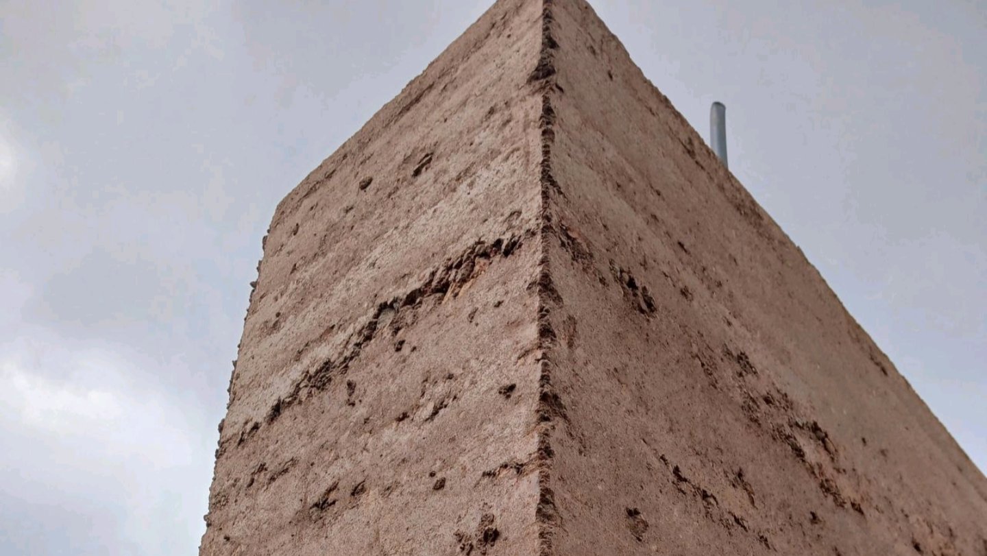 A detailed low-angle close-up view of a tall, modern stabilized rammed earth wall structure showing distinct horizontal soil layers under a cloudy sky, referencing file 249670.png.