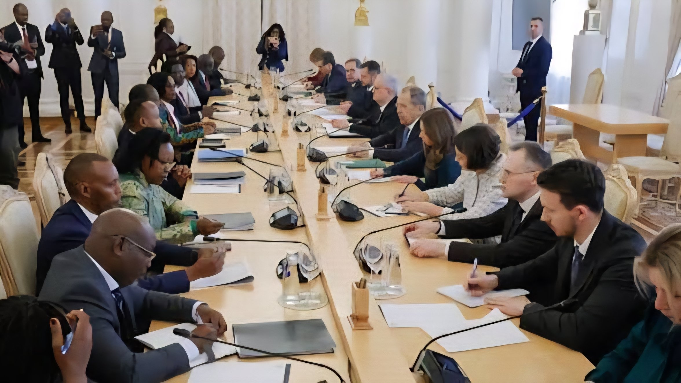 A wide view of a high-level diplomatic meeting around a long wooden table in a formal hall.