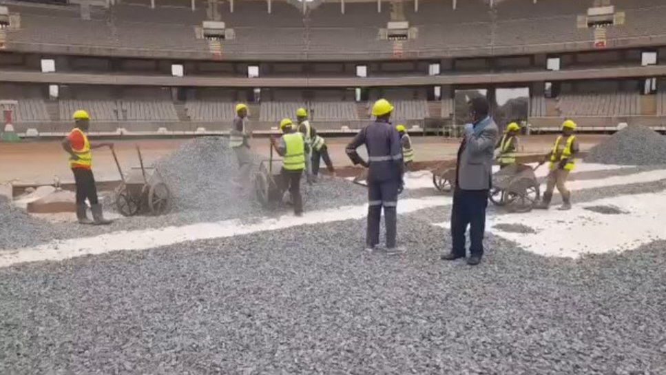 Construction workers distributing stone ballast over a white geotextile membrane on the Kasarani Stadium pitch layer.