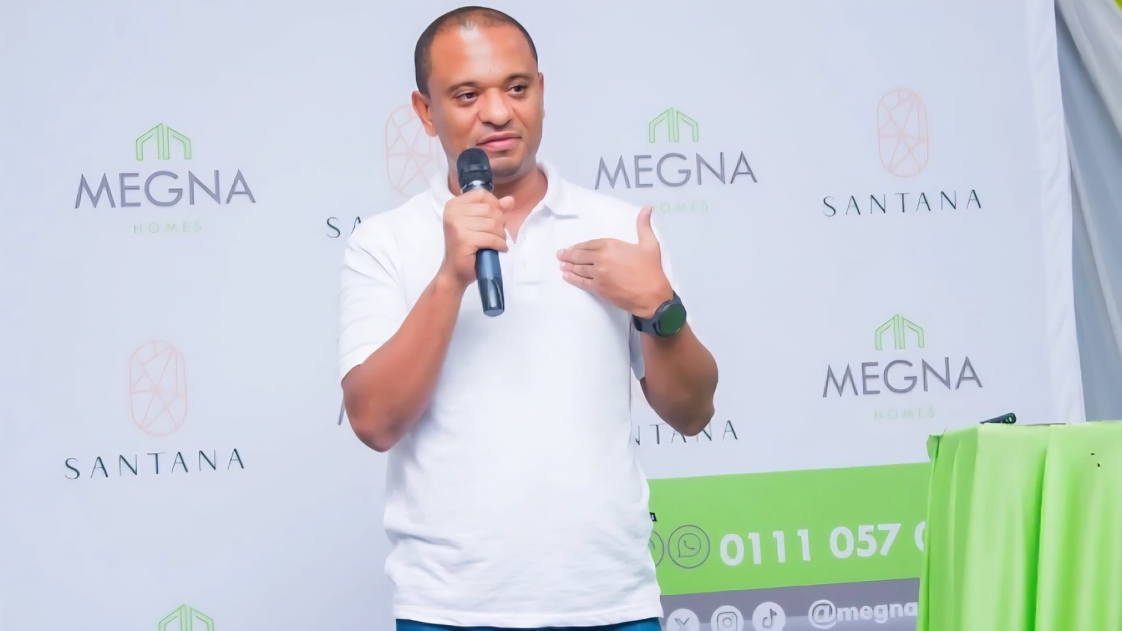 A medium shot of Ahmed Badawy, CEO of Megna Homes, speaking into a microphone during a corporate event with Megna and Santana branding in the background.