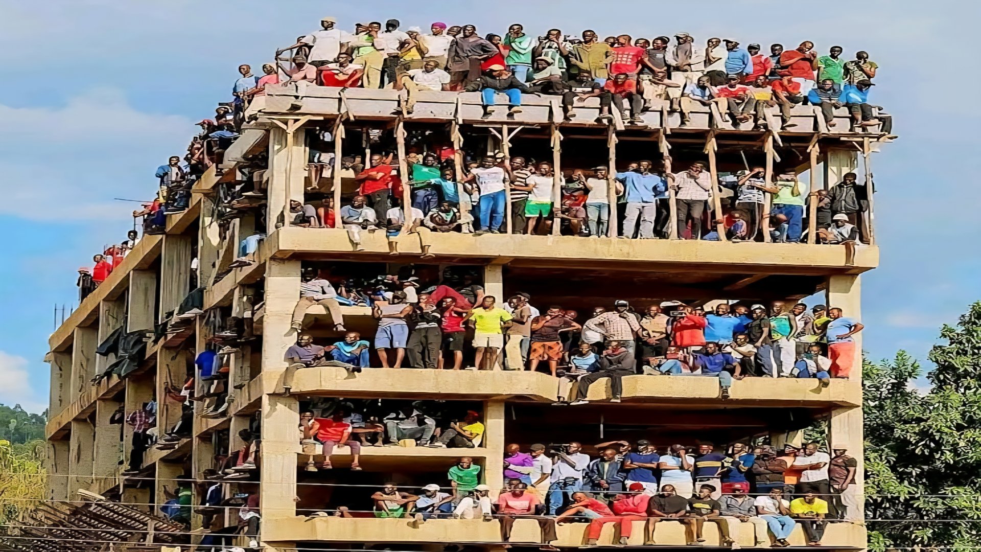 Wide shot of an unfinished four-storey concrete building labeled Milimani Annex with hundreds of people standing and sitting on every floor during a public event.
