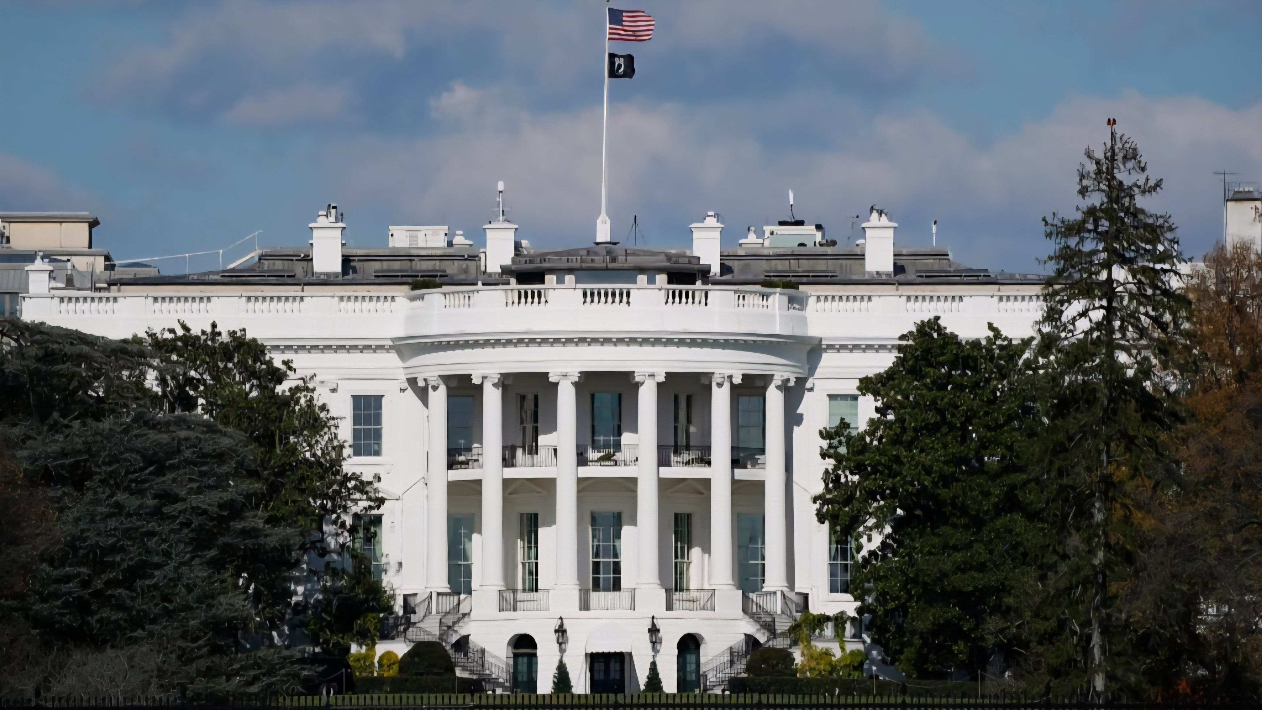 The White House building in Washington, D.C., under a blue sky with the American flag flying at half-staff.