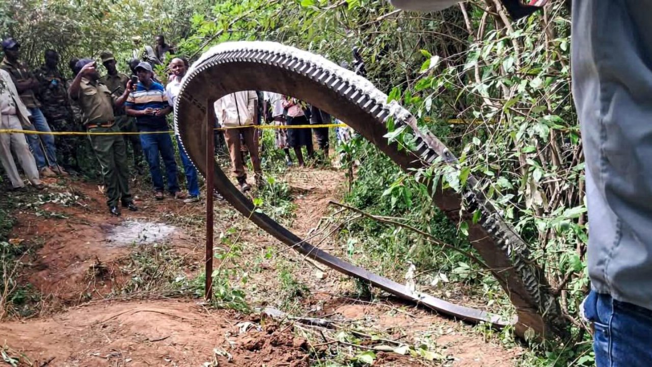 A large, curved metallic ring rests on a dirt path in a wooded area in Kenya, surrounded by local residents and officials behind a security cord.