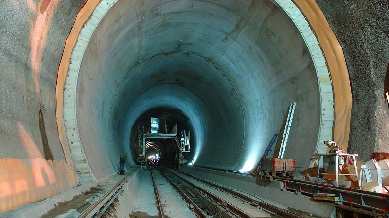 A large industrial construction site at the base of a Swiss mountain showing the portal of a new road tunnel with heavy machinery and equipment.