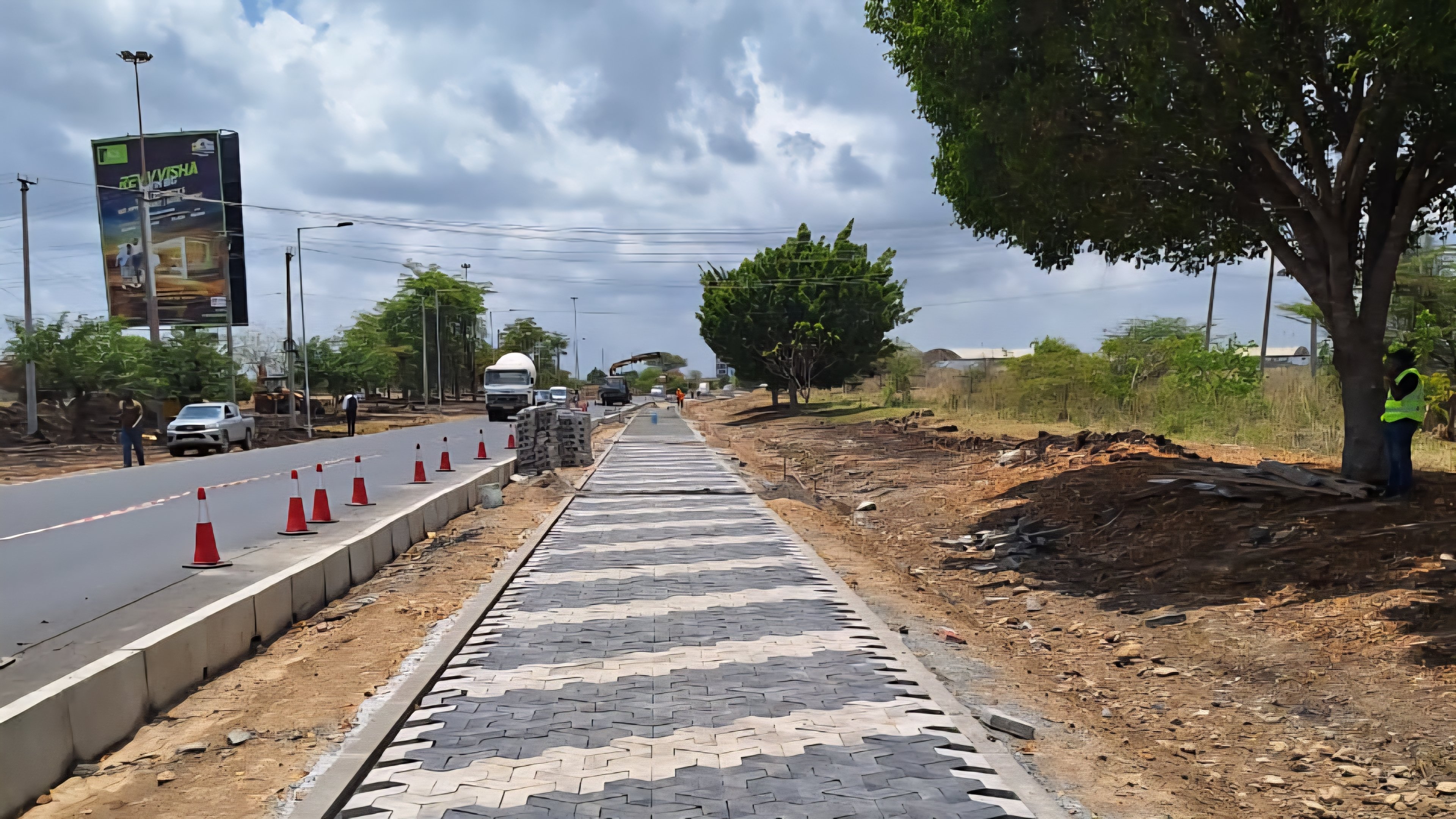 Construction workers wearing high-visibility vests and helmets installing paving blocks for a new pedestrian walkway near the Jomo Kenyatta International Airport entrance.