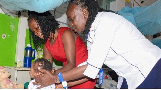 Newborn baby in hospital crib with nurse and parent looking on at Kisumu County Referral Hospita