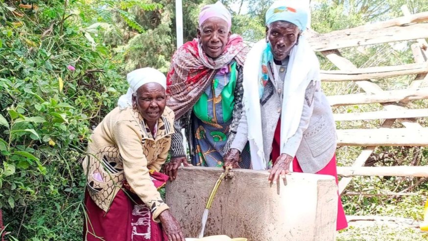 Three elderly women in Bomet stand at a concrete water point, smiling as clean water flows from a tap into a yellow jerrycan.