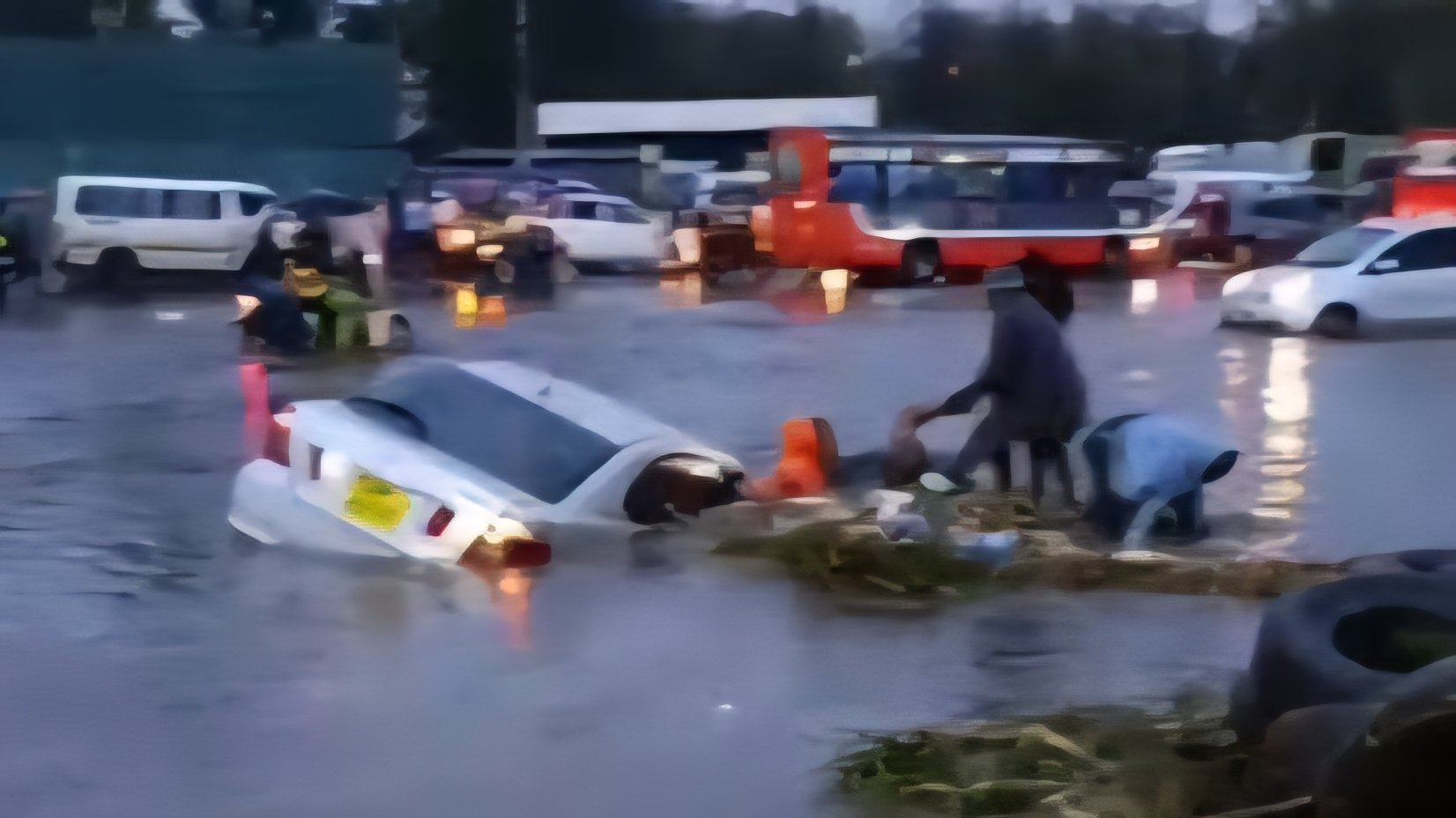 A silver saloon car sits half-submerged in deep floodwater on a flooded road at the Ruai Bypass in Nairobi, with people standing nearby in the rain.