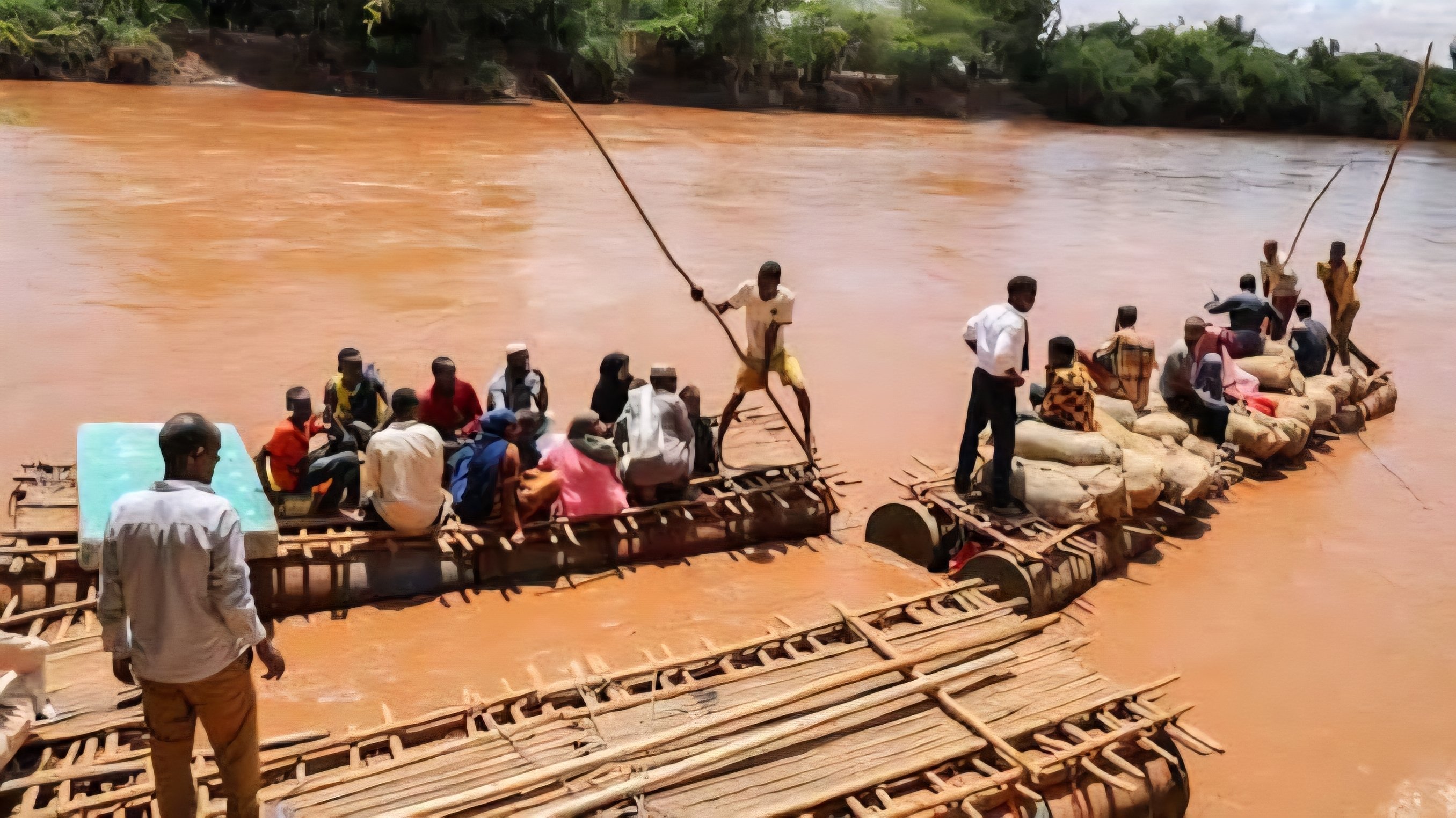 People and cargo on a wooden raft crossing a brown, silt-heavy river.
