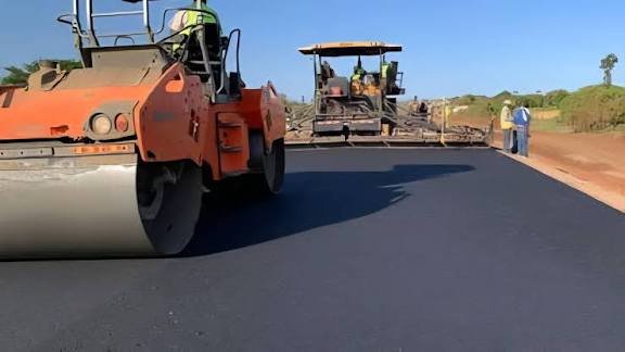 A heavy-duty asphalt paver laying a new road surface in a rural Kenyan landscape with a small cart on the side.