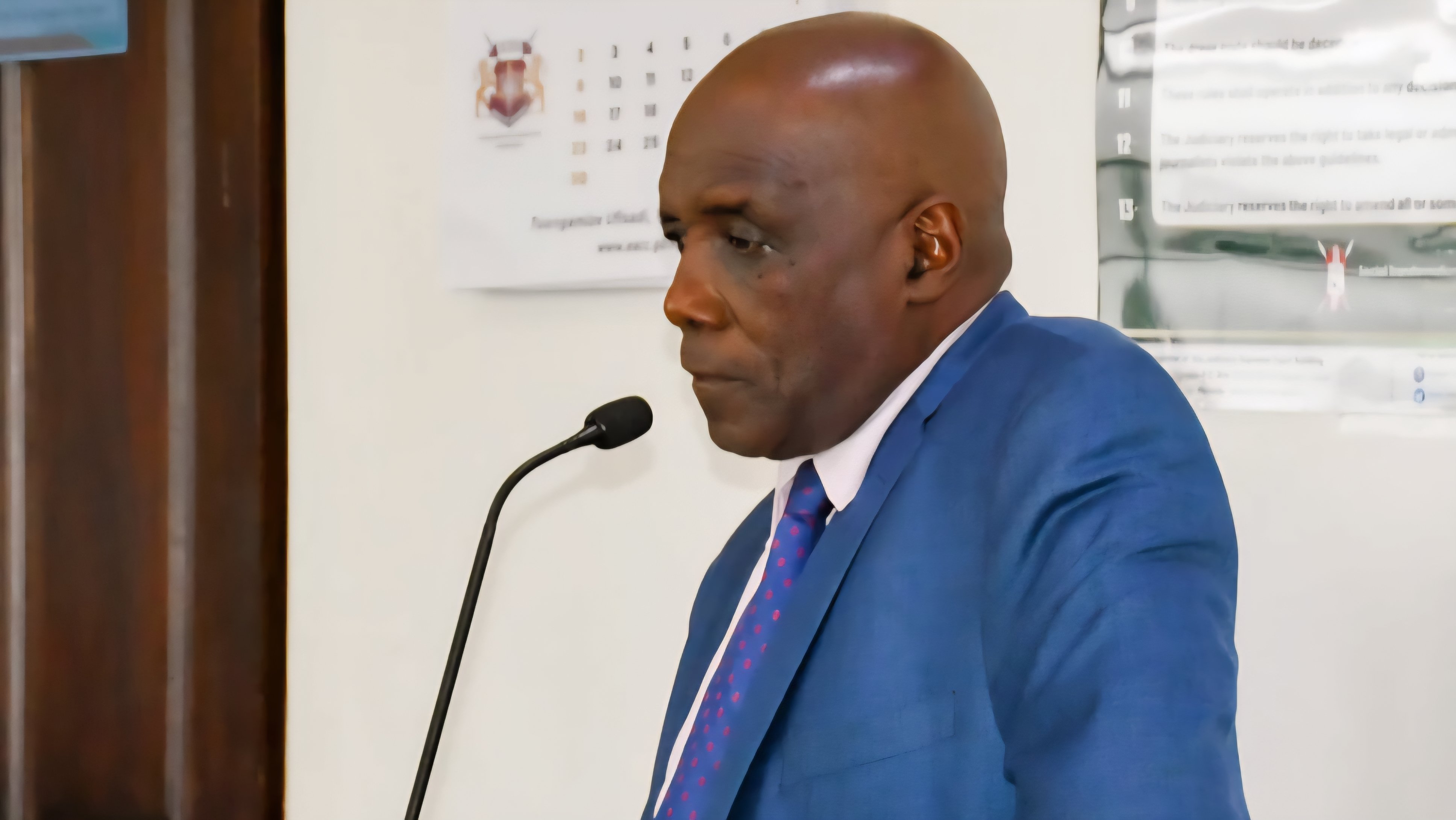 A medium shot of Muhammad Swazuri standing in a courtroom dock during a legal proceeding.