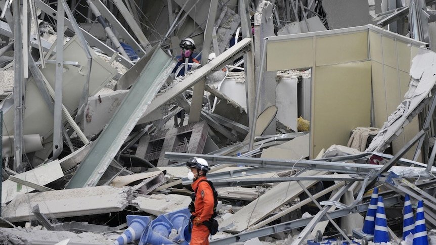 Rescuers in bright protective gear search through the pile of a collapsed house with tiled roofs following an earthquake in Japan.