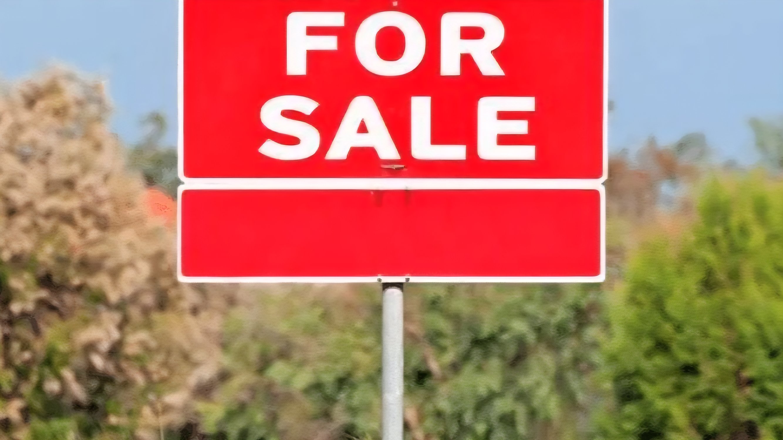 A red 'For Sale' sign standing in front of green trees and a blurred commercial building structure in the background.