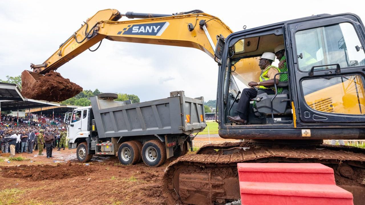 Featured image showing a yellow Sany excavator performing the official groundbreaking at Gusii Stadium in Kisii, with a truck positioned nearby and a large crowd of spectators in the background stands.