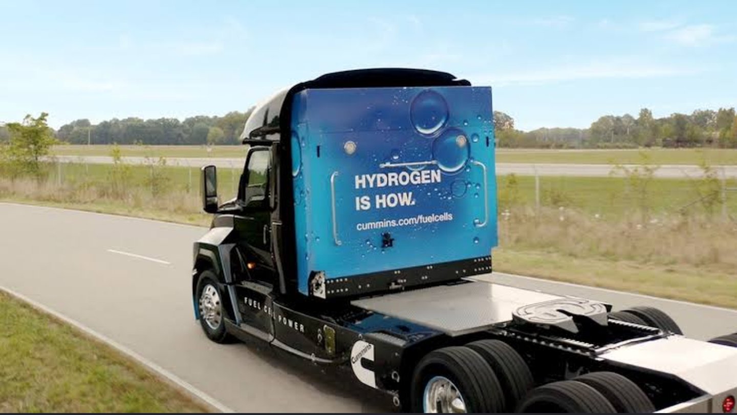 A black heavy-duty truck chassis featuring a prominent blue hydrogen fuel cell unit mounted behind the cab, driving on an open asphalt roadway alongside a green field under a clear sky.