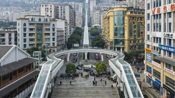 A wide aerial view of a dense, hilly cityscape in China featuring a long, covered escalator structure winding between high-rise buildings and mountain slopes.