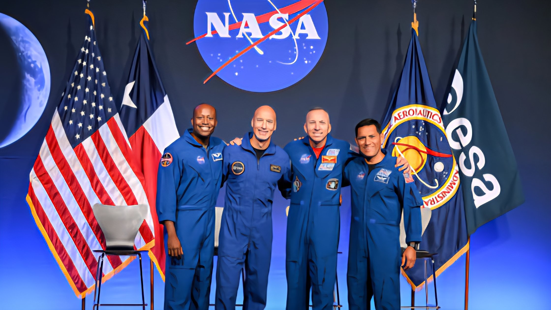 The four male astronauts selected for the NASA Artemis III test mission standing together in blue flight suits in front of the NASA logo and flags.