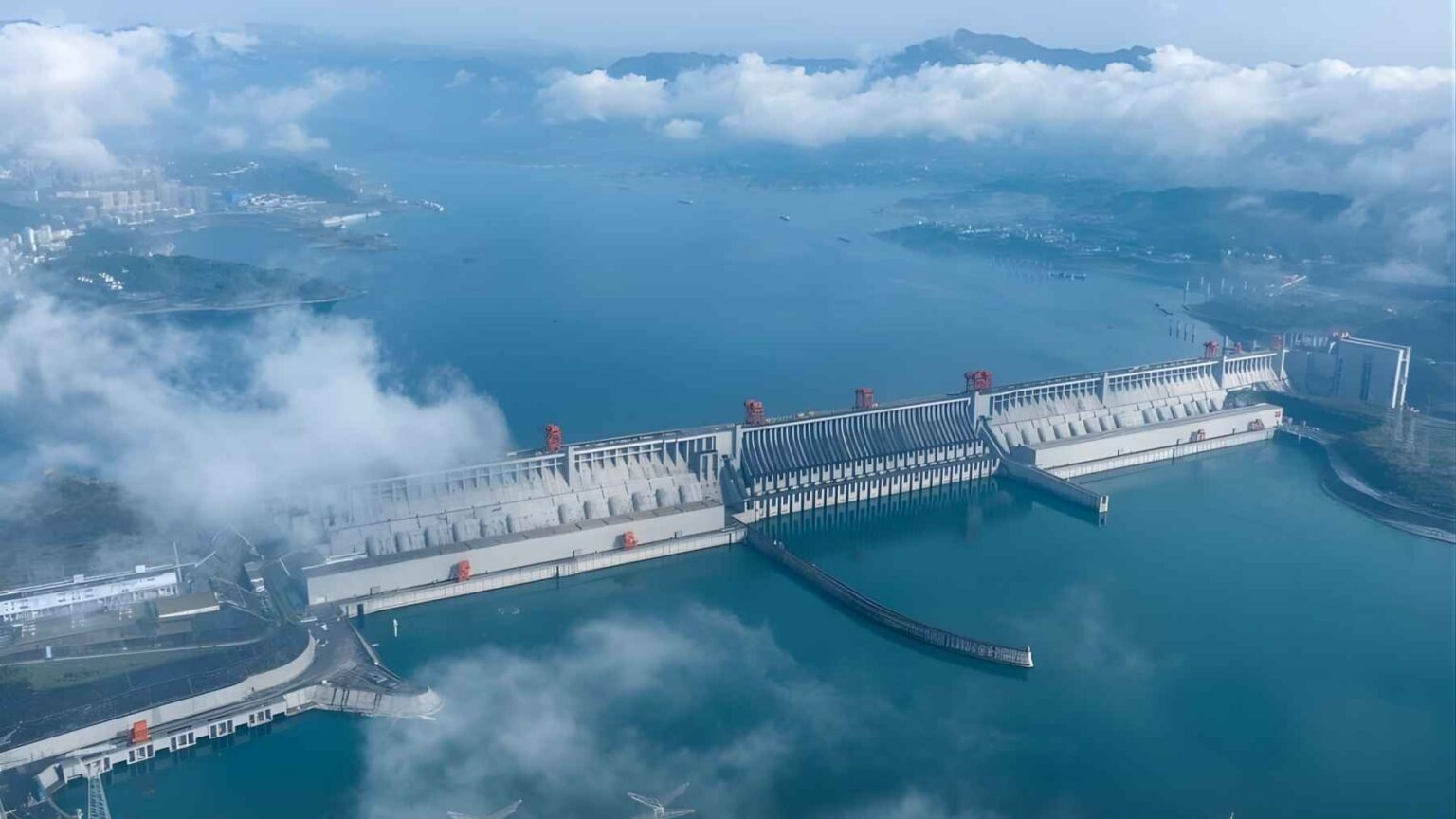 High-angle aerial view of the massive Three Gorges Dam spanning the Yangtze River in China, showing the concrete gravity structure and the vast reservoir behind it under a light mist.