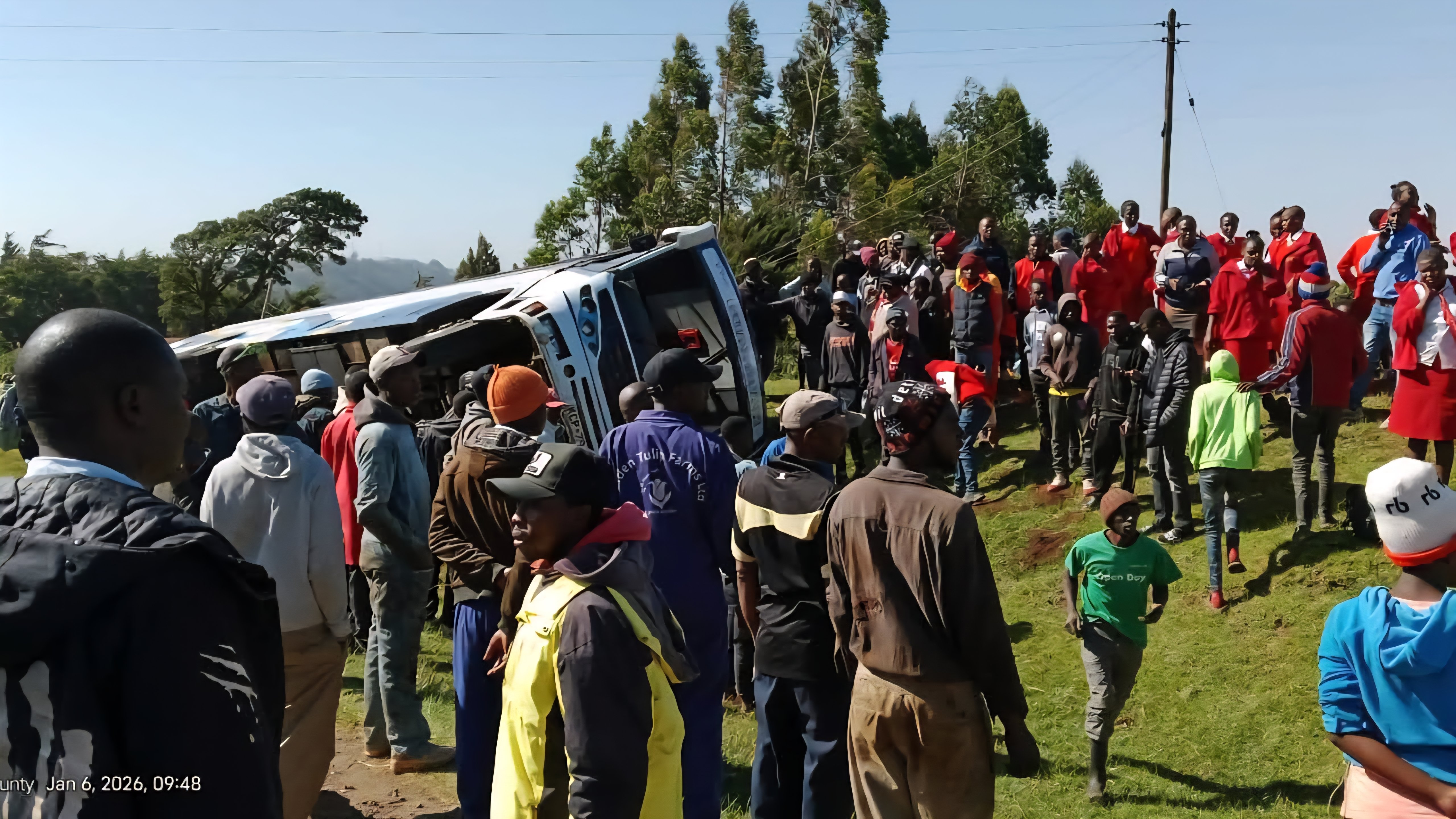 A Guardian Angel public service bus lying on its side off the shoulder of a paved highway surrounded by onlookers and emergency responders.