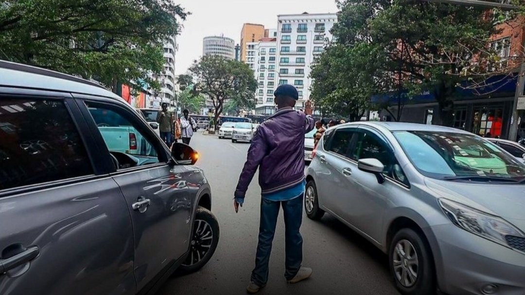 A man in a purple jacket directing a silver car into a parking spot on a busy Nairobi street.