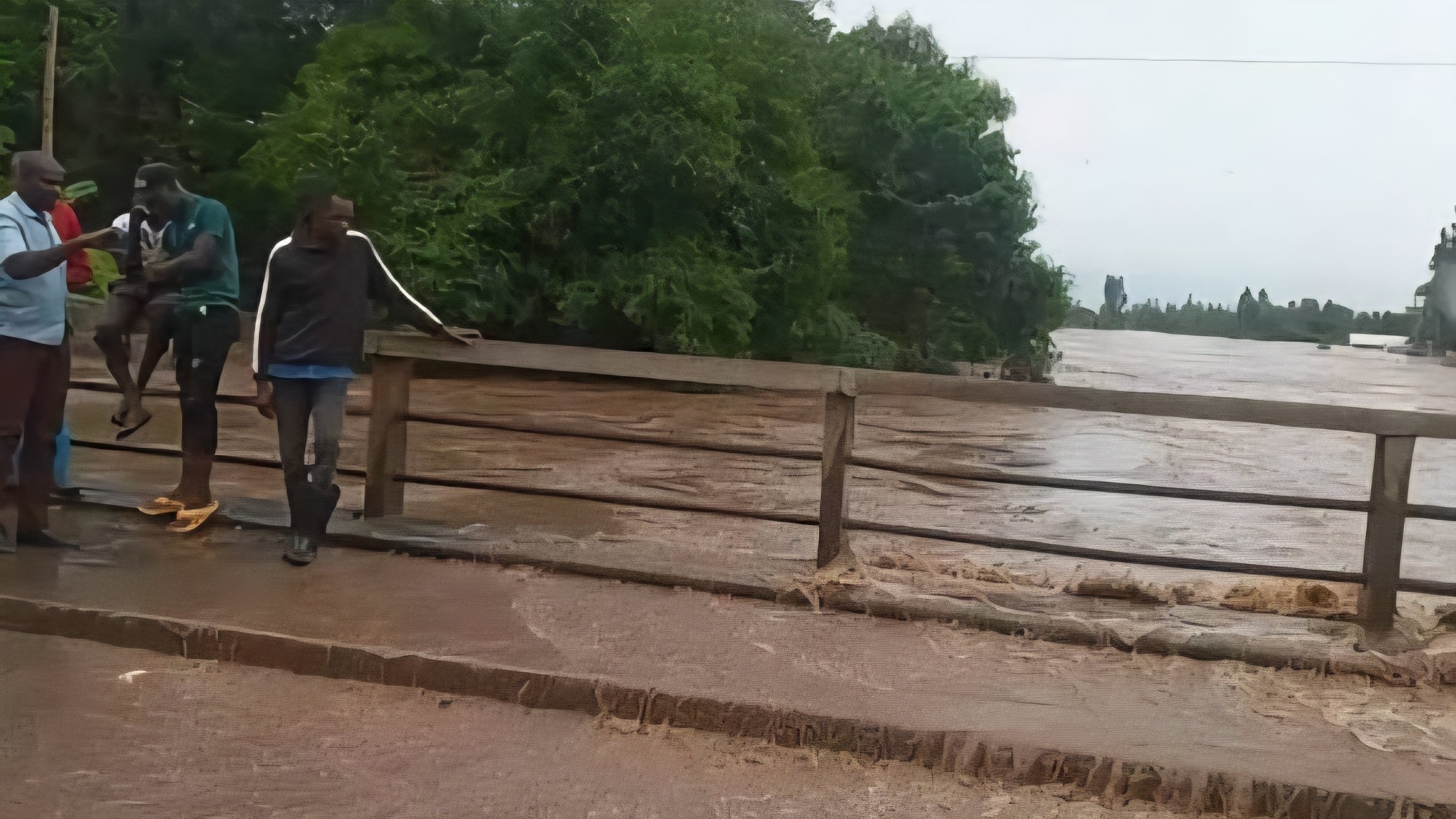 Turbulent brown floodwaters from River Nyando overflowing the Ahero Bridge and submerging sections of the asphalt highway, with no vehicles visible on the flooded stretch.