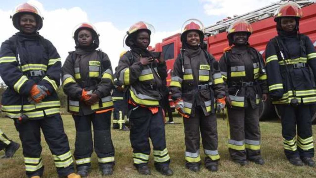 Six female firefighters wearing black protective uniforms and yellow helmets stand side-by-side in front of a red fire engine.