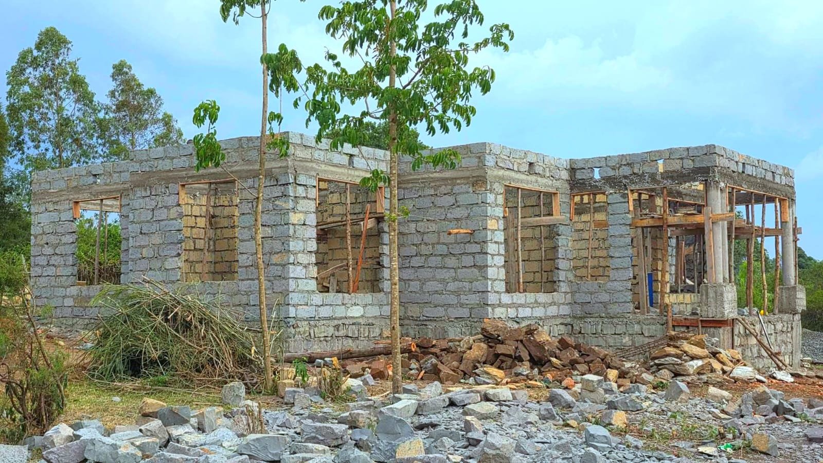 A residential building under construction in Kenya featuring stone masonry walls and large openings for framed corner windows.