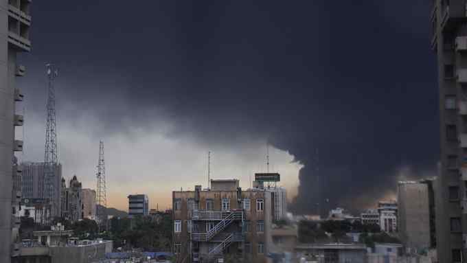 Thick black smoke billows from a dense urban skyline following an explosion in a city under military conflict.
