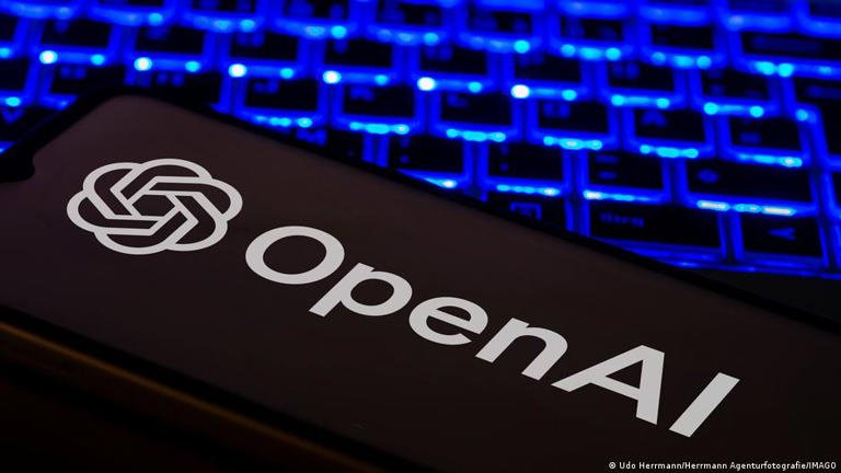 A close-up view of a computer keyboard illuminated in blue light displaying the OpenAI logo on an adjacent mobile screen.
