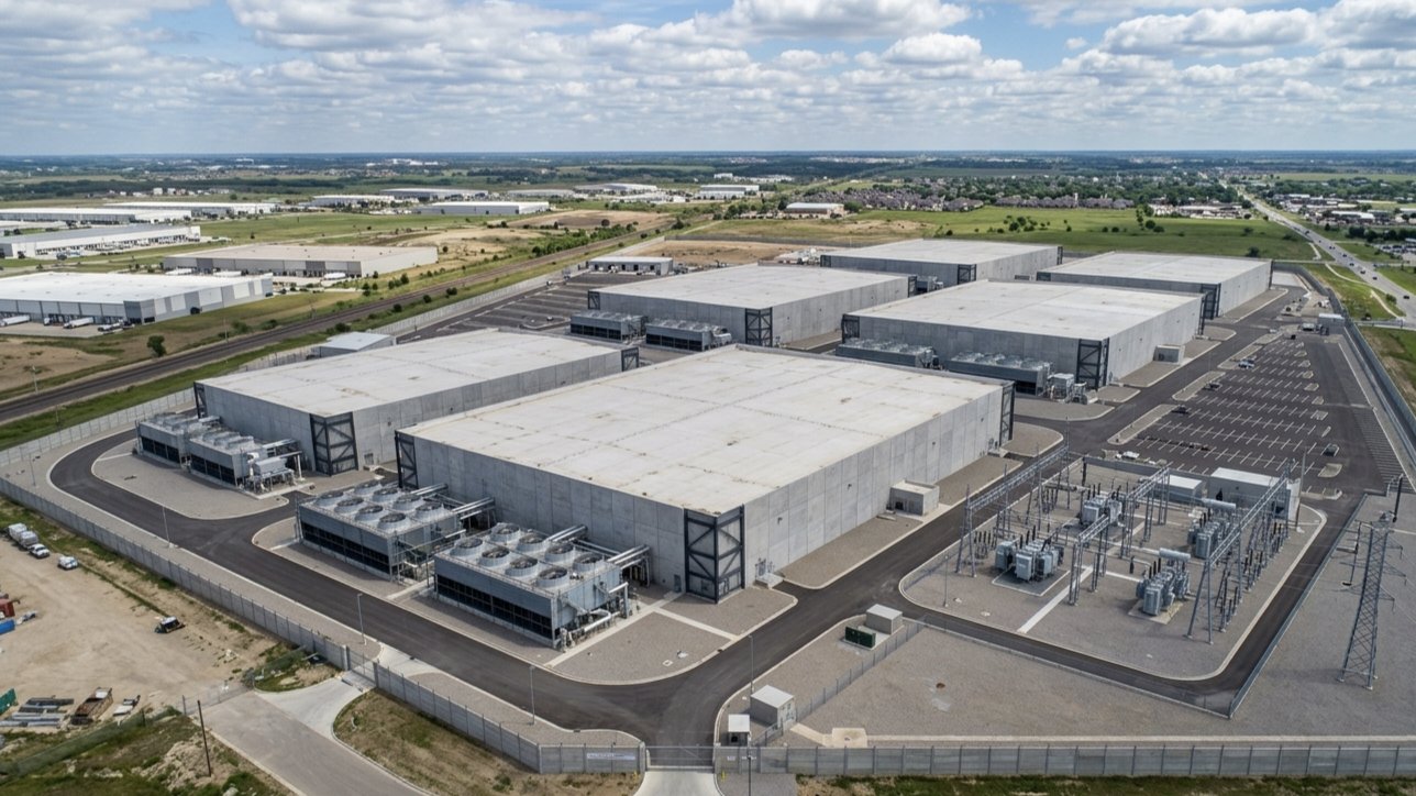 Aerial view of a large hyperscale data center campus showing multiple windowless server buildings, cooling infrastructure and power substations on an industrial site.