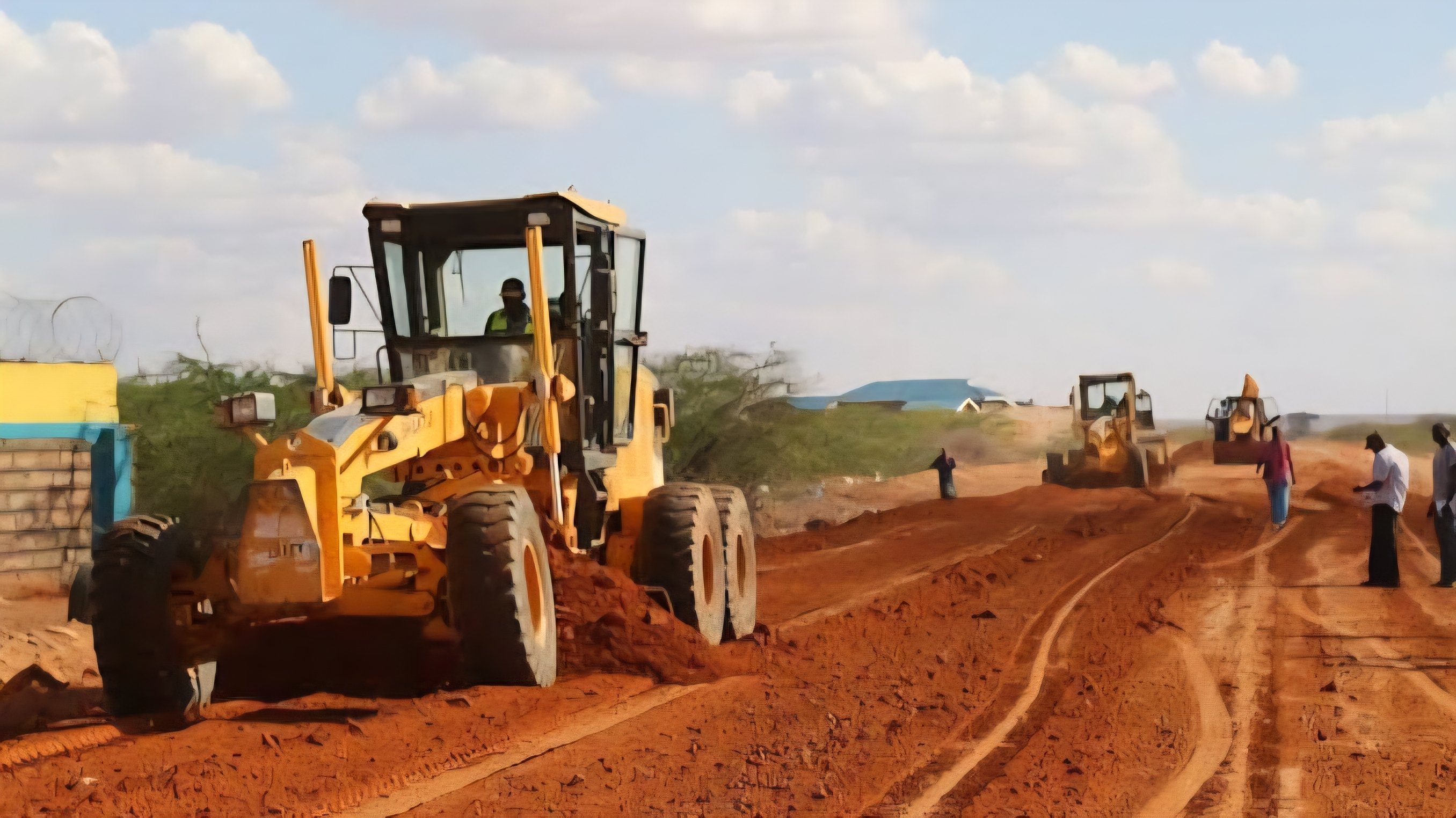 A motor grader and a soil compactor working on a wide, cleared earth section of the Lamu-Ijara-Garissa road under a clear sky.