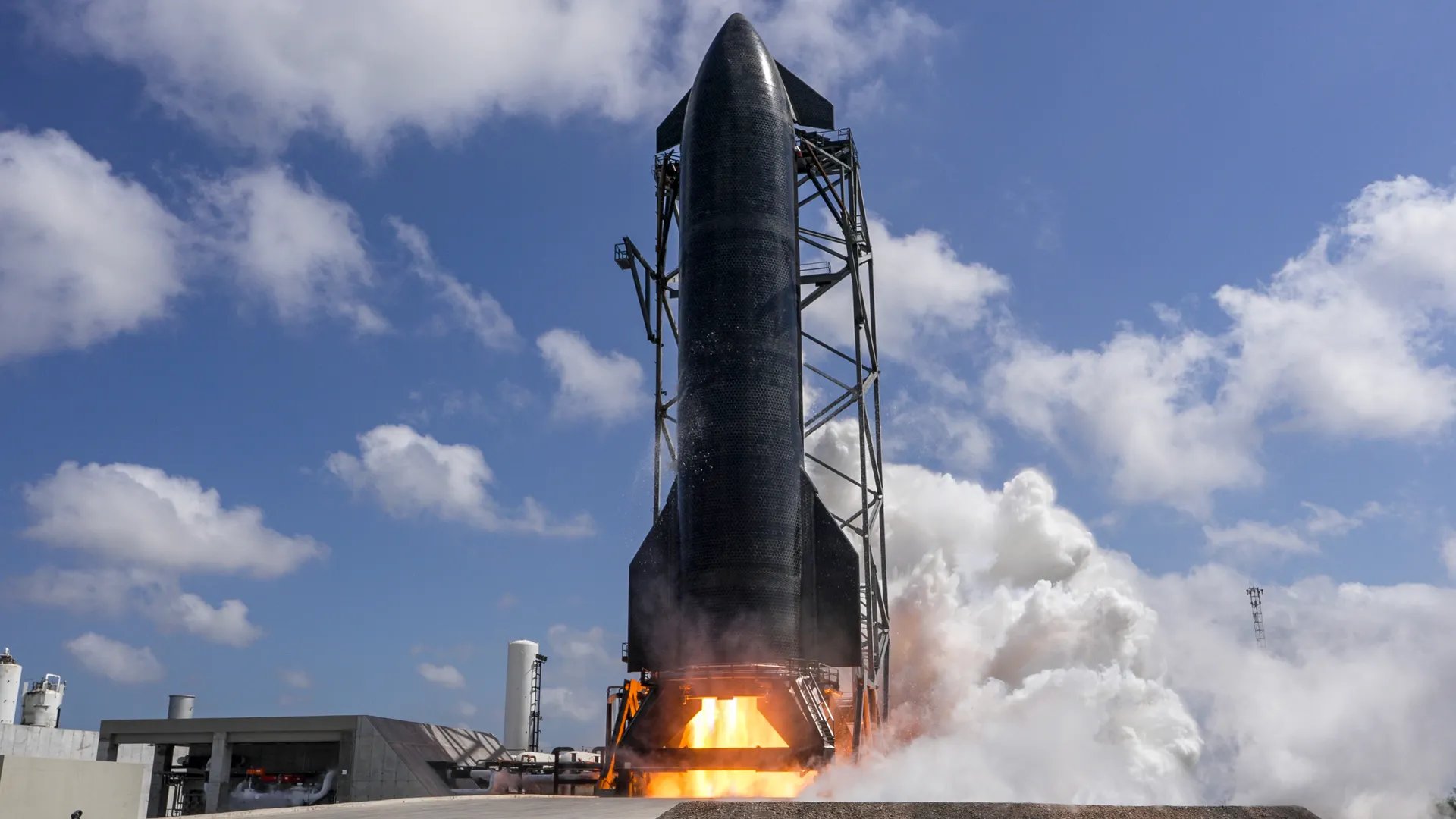 A large stainless-steel rocket stage covered in black thermal tiles stands on a test mount during a static fire test with flames emitting from its base.