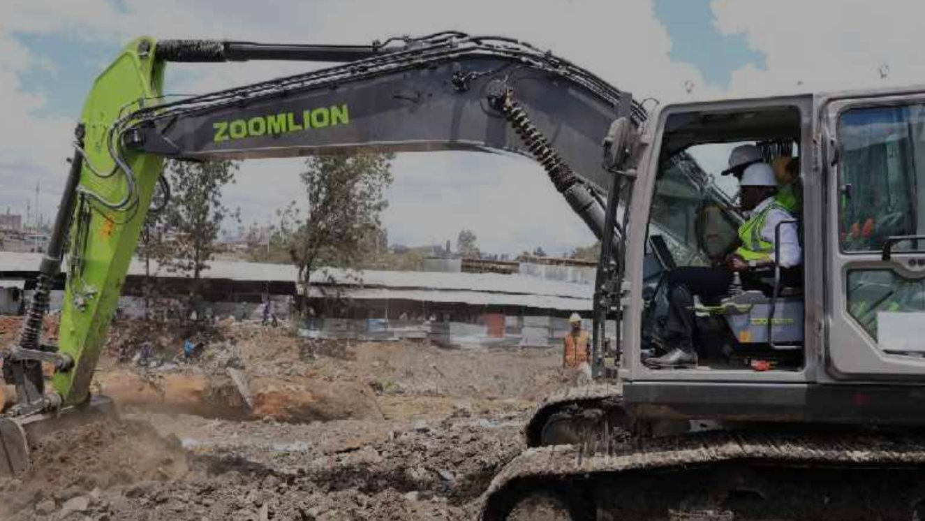 A white and lime green Zoomlion excavator sits on a dusty construction site in Gikomba market with workers in high-visibility vests nearby.