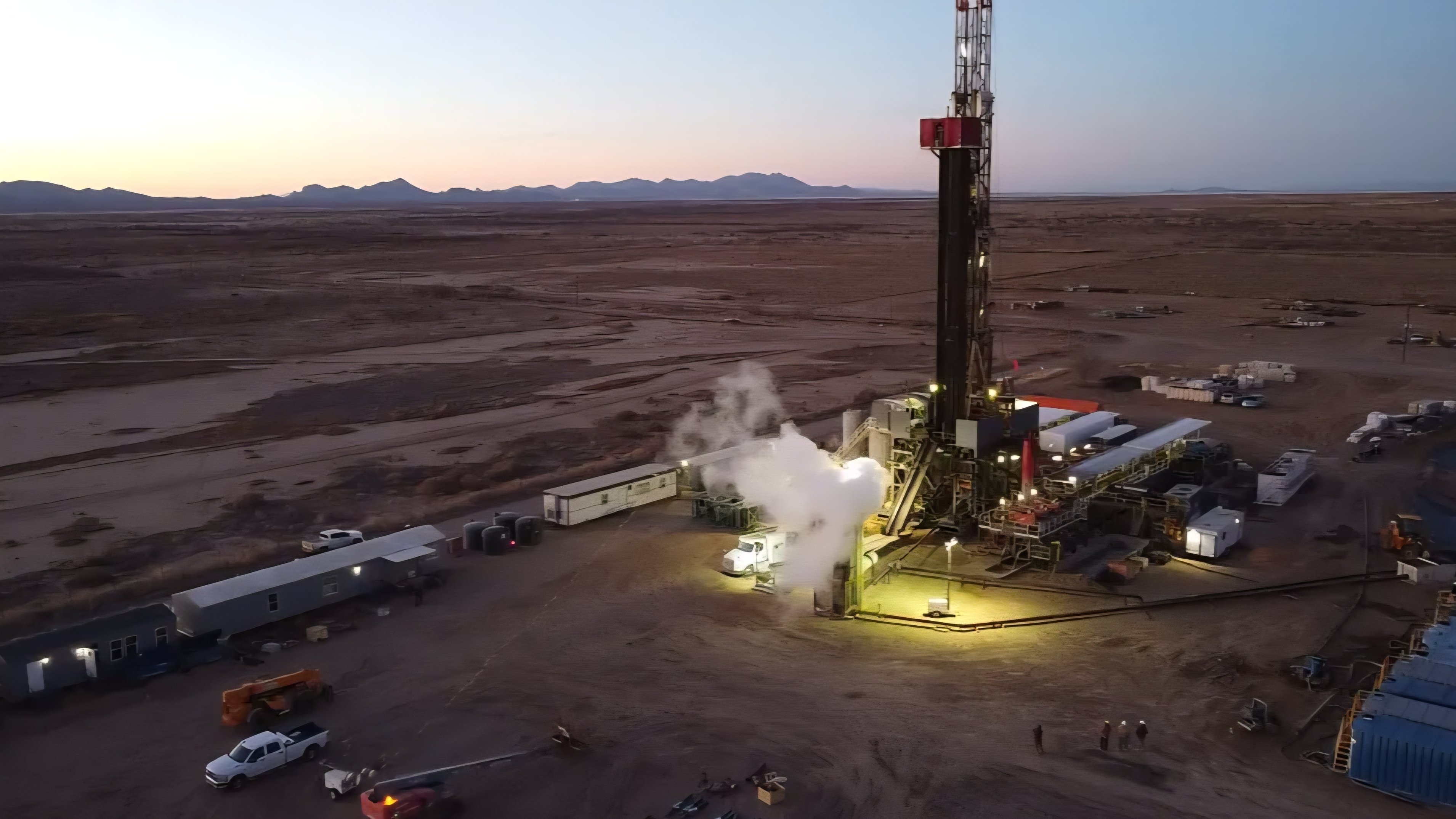 Aerial view at dusk of a geothermal drilling rig and surface power facility operating in an arid desert terrain.