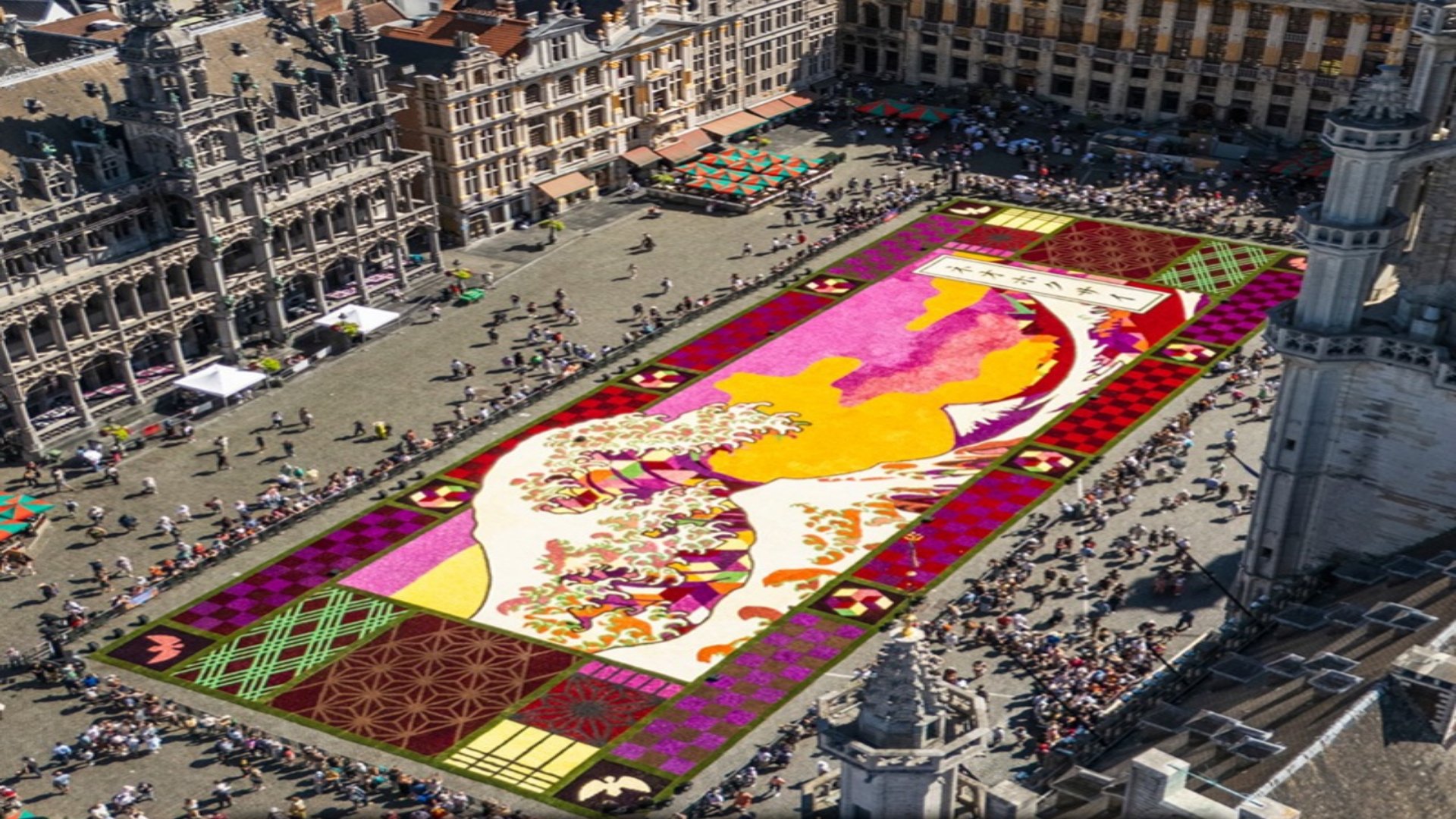 Aerial view of the 2026 Brussels Flower Carpet laid out across the paved Grand-Place, featuring a floral design of Hokusai's iconic wave artwork surrounded by historic architecture.