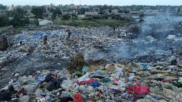 Wide view of a massive, smoldering informal dumpsite in Kitengela, Kenya, showing layers of plastic waste and smoke rising against a backdrop of distant residential buildings.