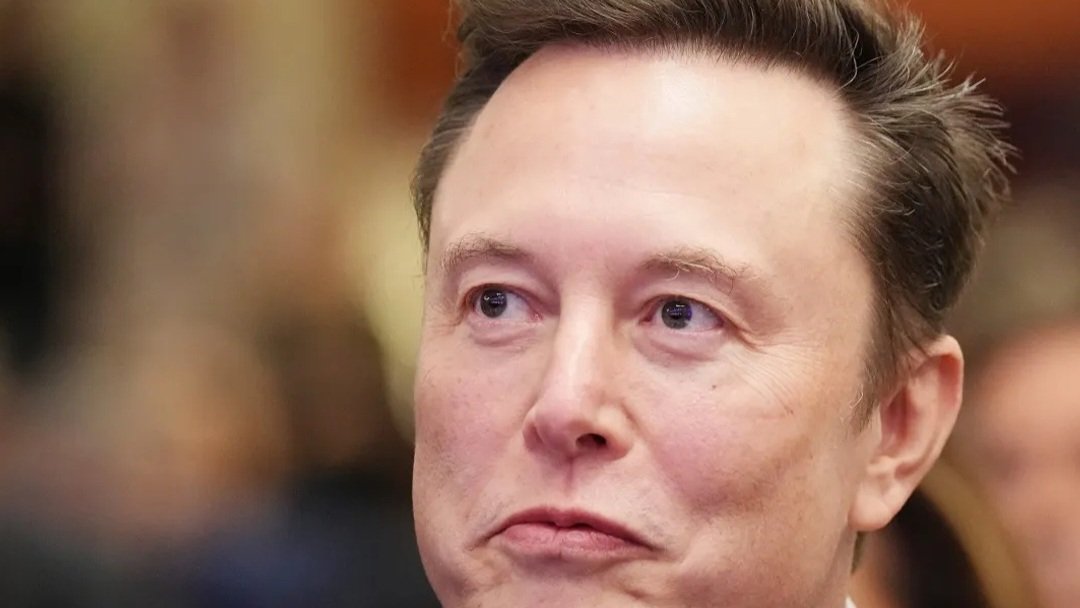 Close-up profile of Elon Musk in a dark suit and tie looking forward with a serious expression.